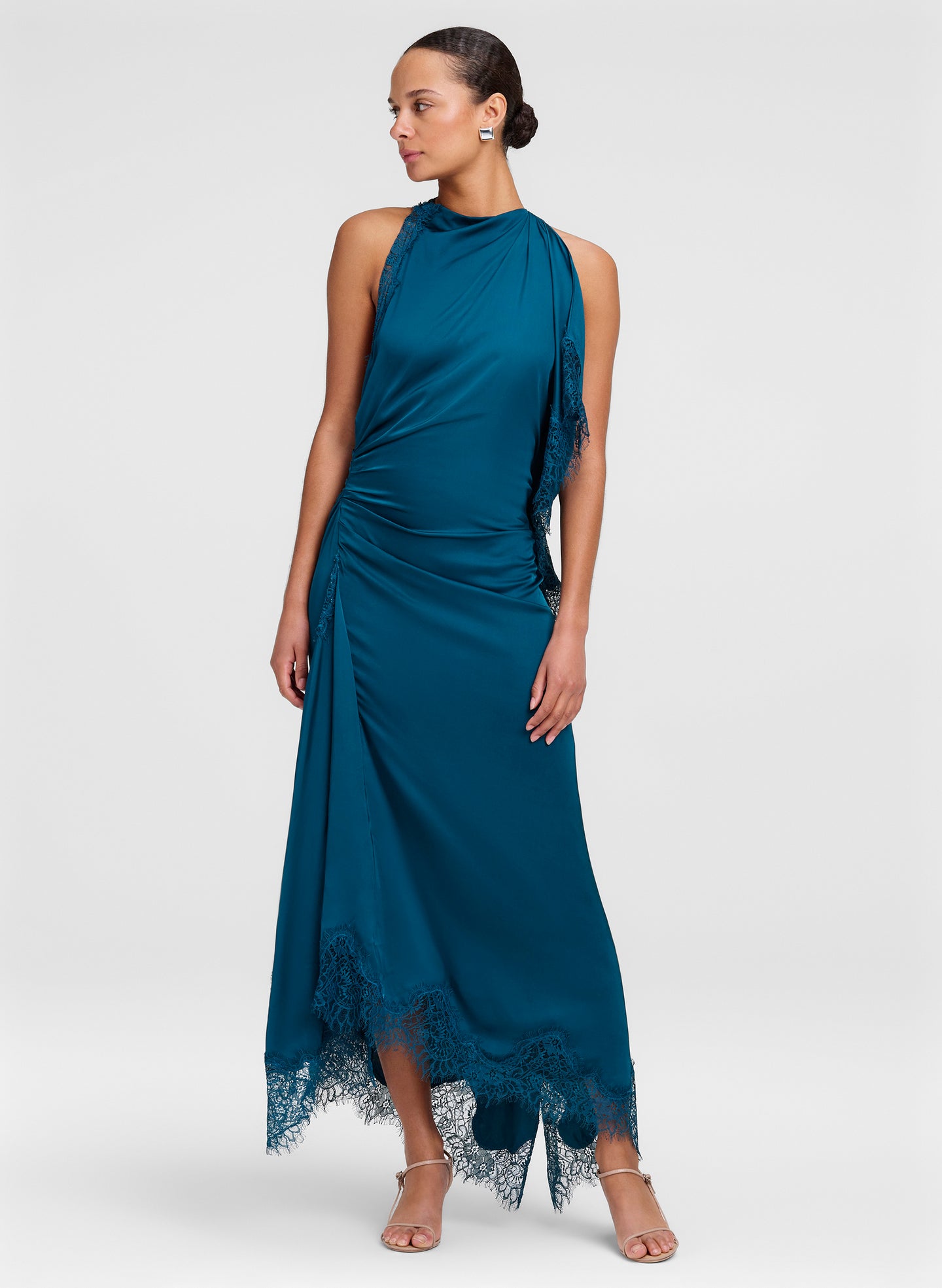 A woman modeling a sleeveless teal blue maxi dress with a draped halter-style neckline, gathered skirt, and lace trim at the hem, photographed against a light gray studio background.