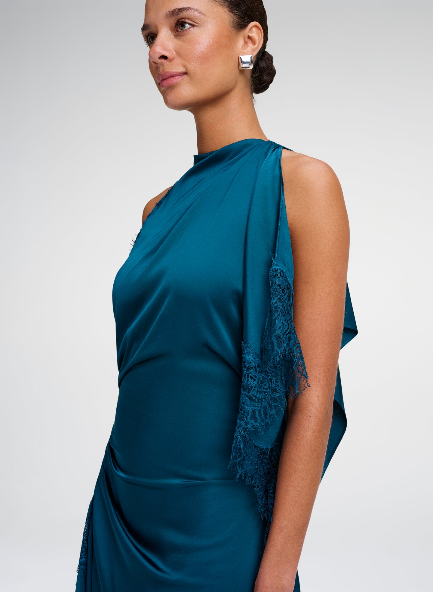 A woman wearing a teal blue satin dress with a draped lace-trimmed shawl over one shoulder, shown in a side profile against a light gray studio background.