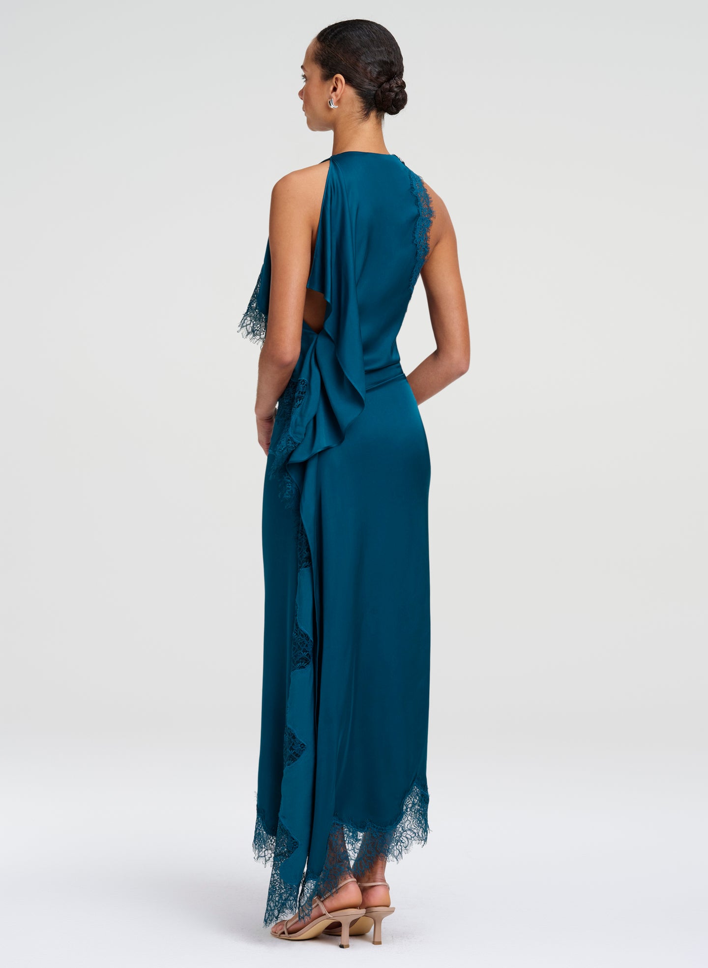 A woman modeling a teal blue one-shoulder maxi dress from the back, featuring a draped halter-style neckline, sheer side cutouts, and lace trim at the hem, set against a plain light gray studio background.