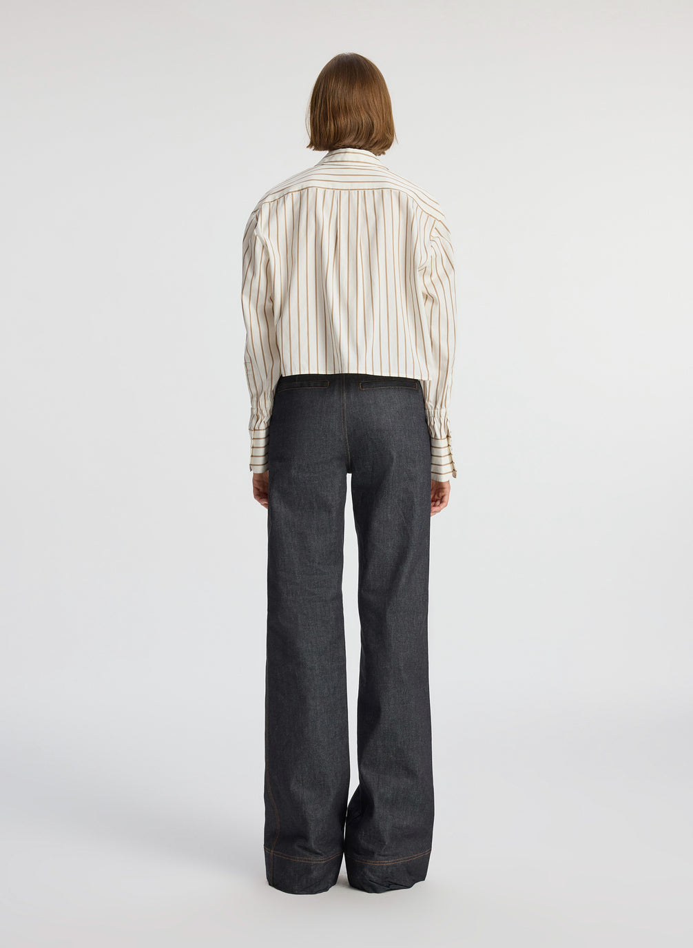 back view of woman wearing cream and brown striped collared shirt and dark wash raw denim jeans