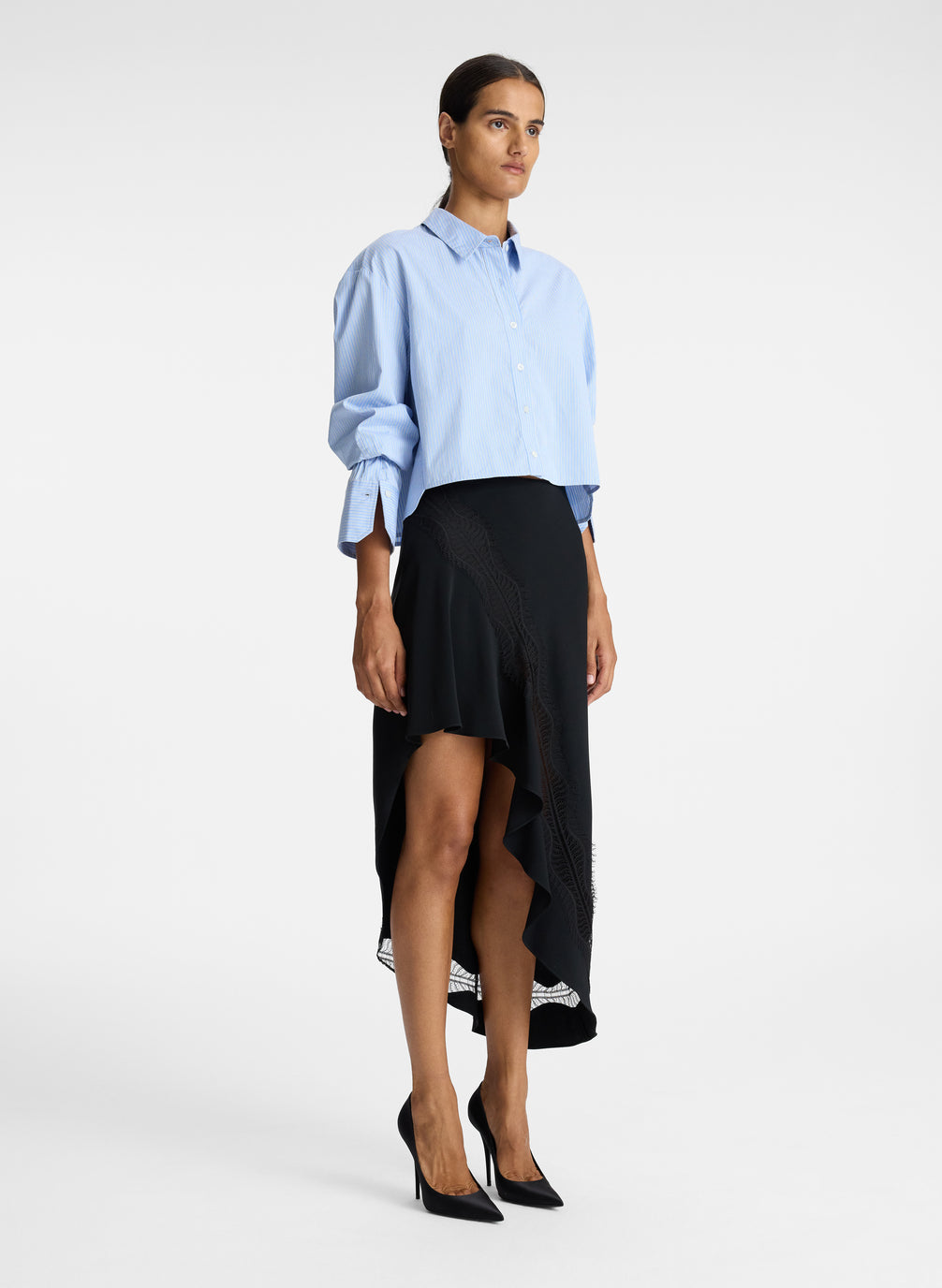 side view of woman wearing light blue striped oversized cropped button down shirt and black asymmetric midi skirt