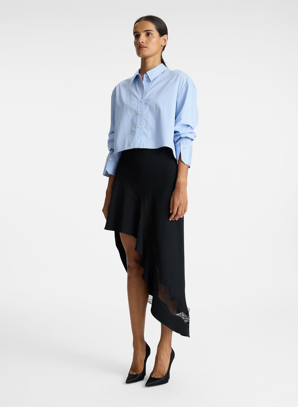 side view of woman wearing blue striped button down collared shirt and asymmetric black satin and lace skirt