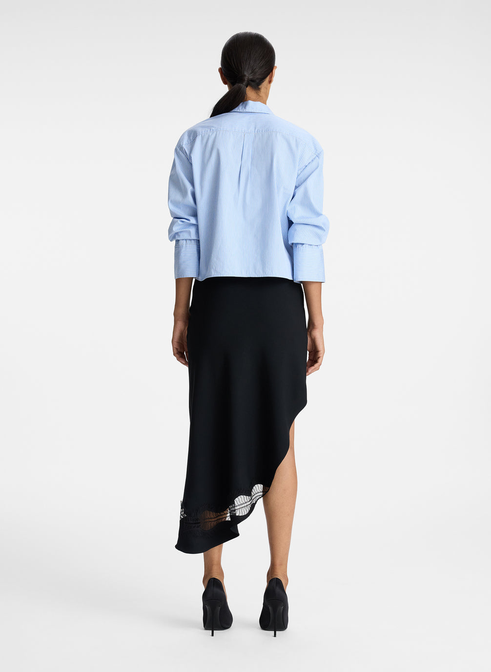 back view of woman wearing light blue striped oversized cropped button down shirt and black asymmetric midi skirt