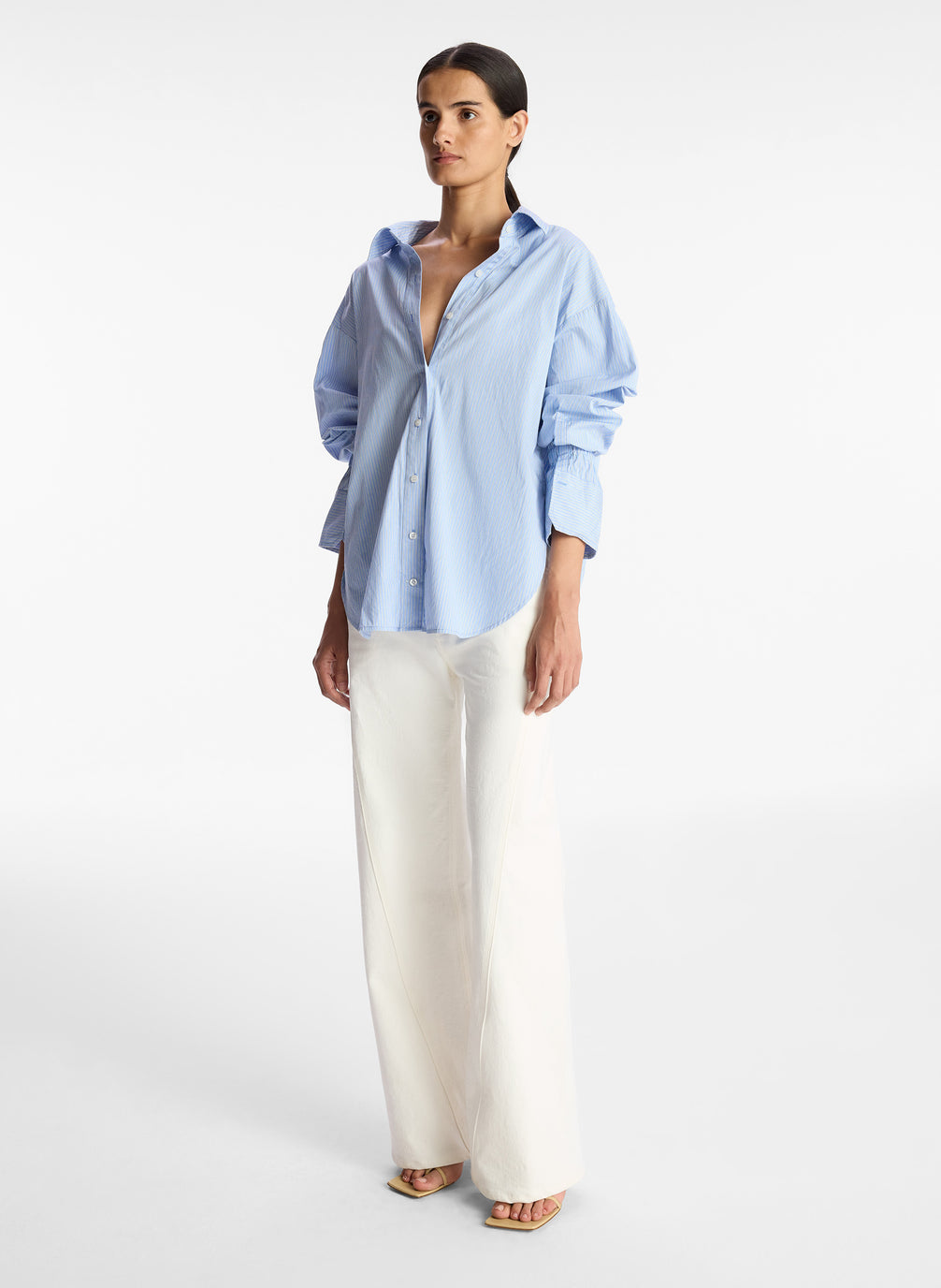side view of woman wearing light blue striped oversized button down shirt and white pants