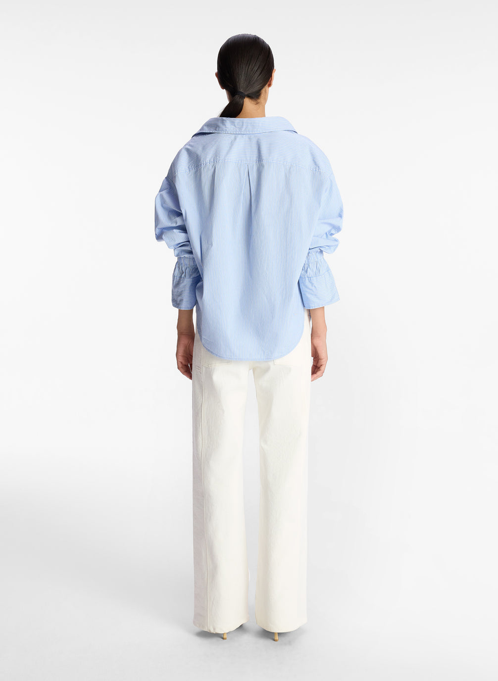 back view of woman wearing light blue striped oversized button down shirt and white pants