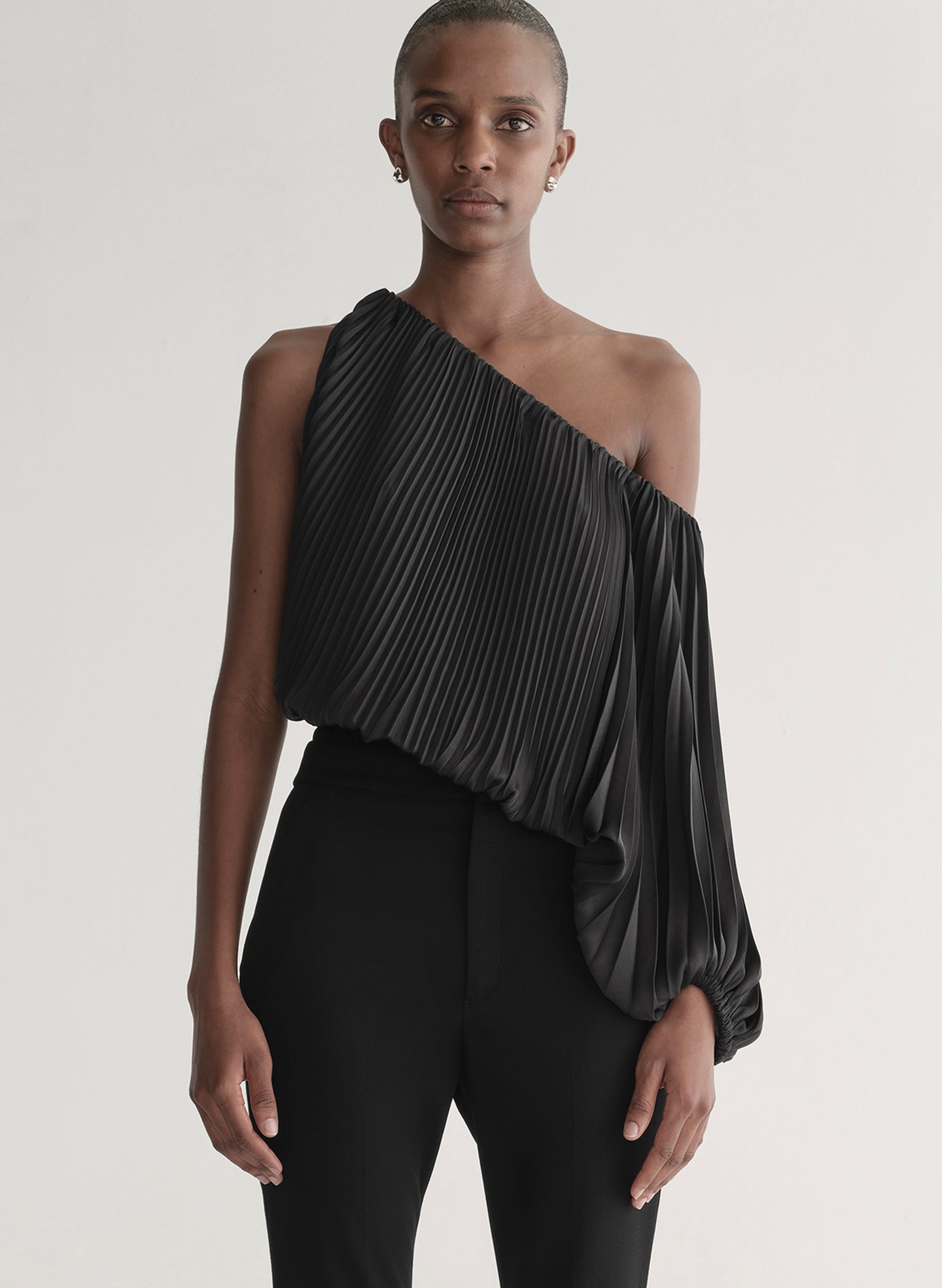 Woman wearing a black pleated one-shoulder top and black pants against a plain background