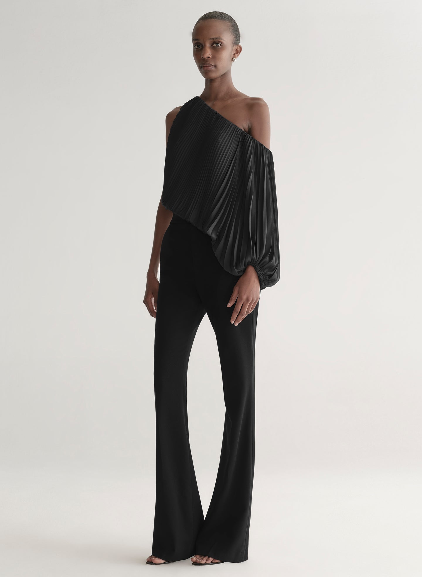 Woman wearing a black pleated one-shoulder top and black pants against a plain background
