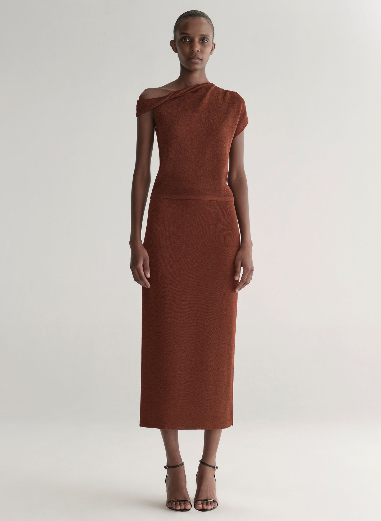 Woman wearing a rust one shoulder knit top and matching skirt against a plain background