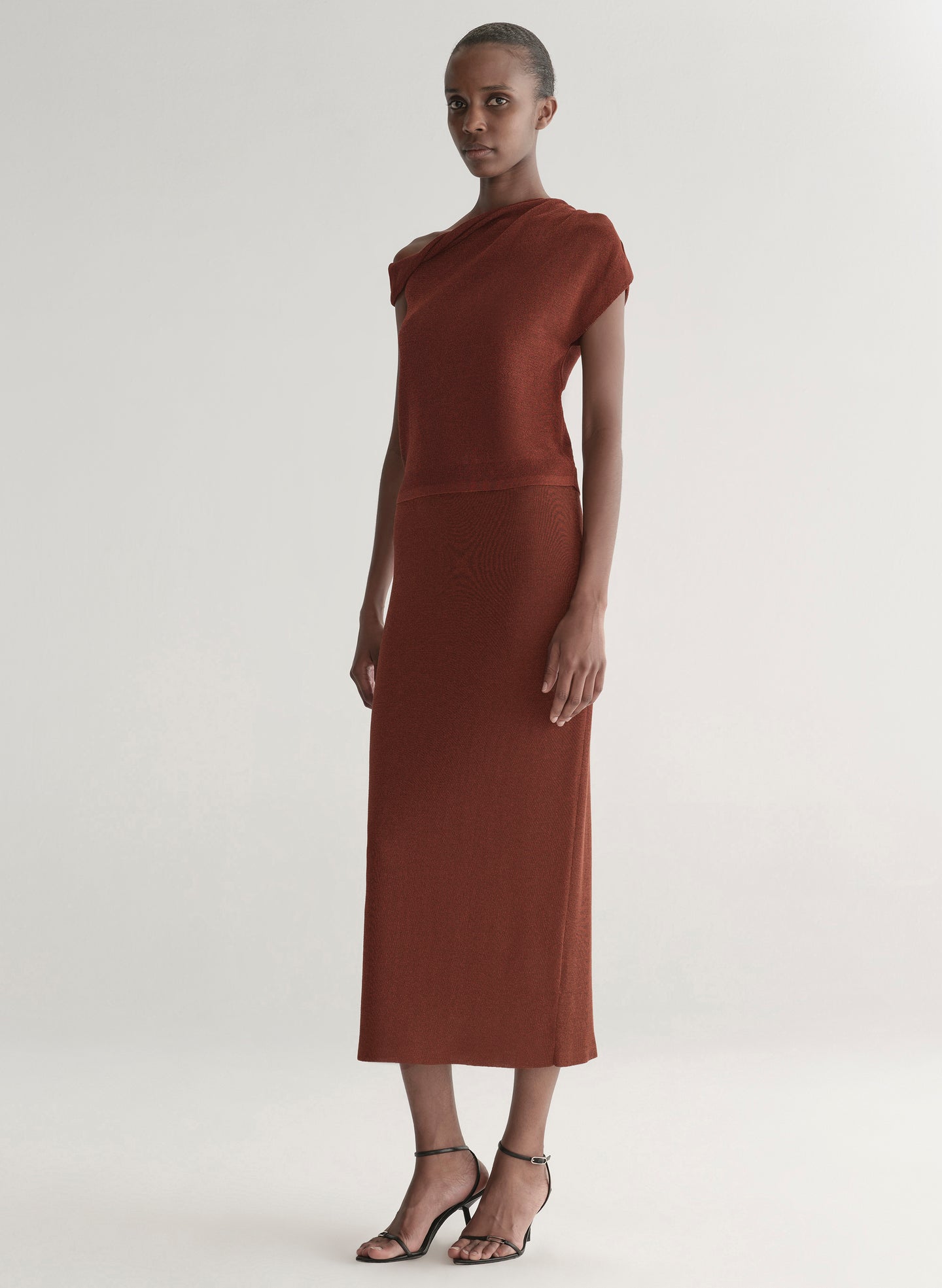 Woman wearing a rust one shoulder knit top and matching skirt against a plain background