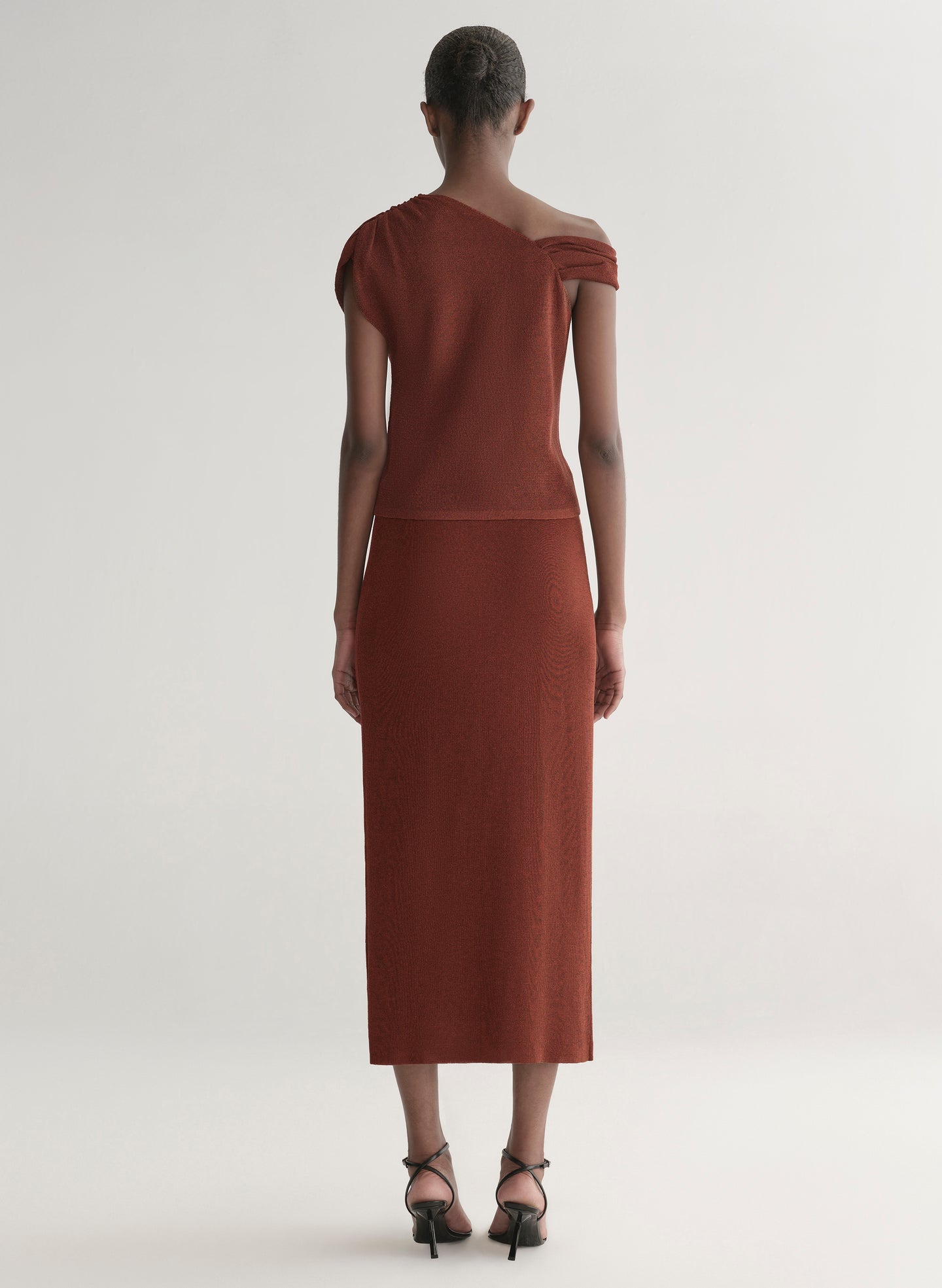 Woman wearing a rust one shoulder knit top and matching skirt against a plain background