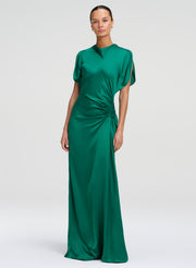 A woman modeling a floor-length emerald green satin gown with short flutter sleeves, a high neckline, and a gathered ruched detail at the waist, photographed against a light gray studio background.