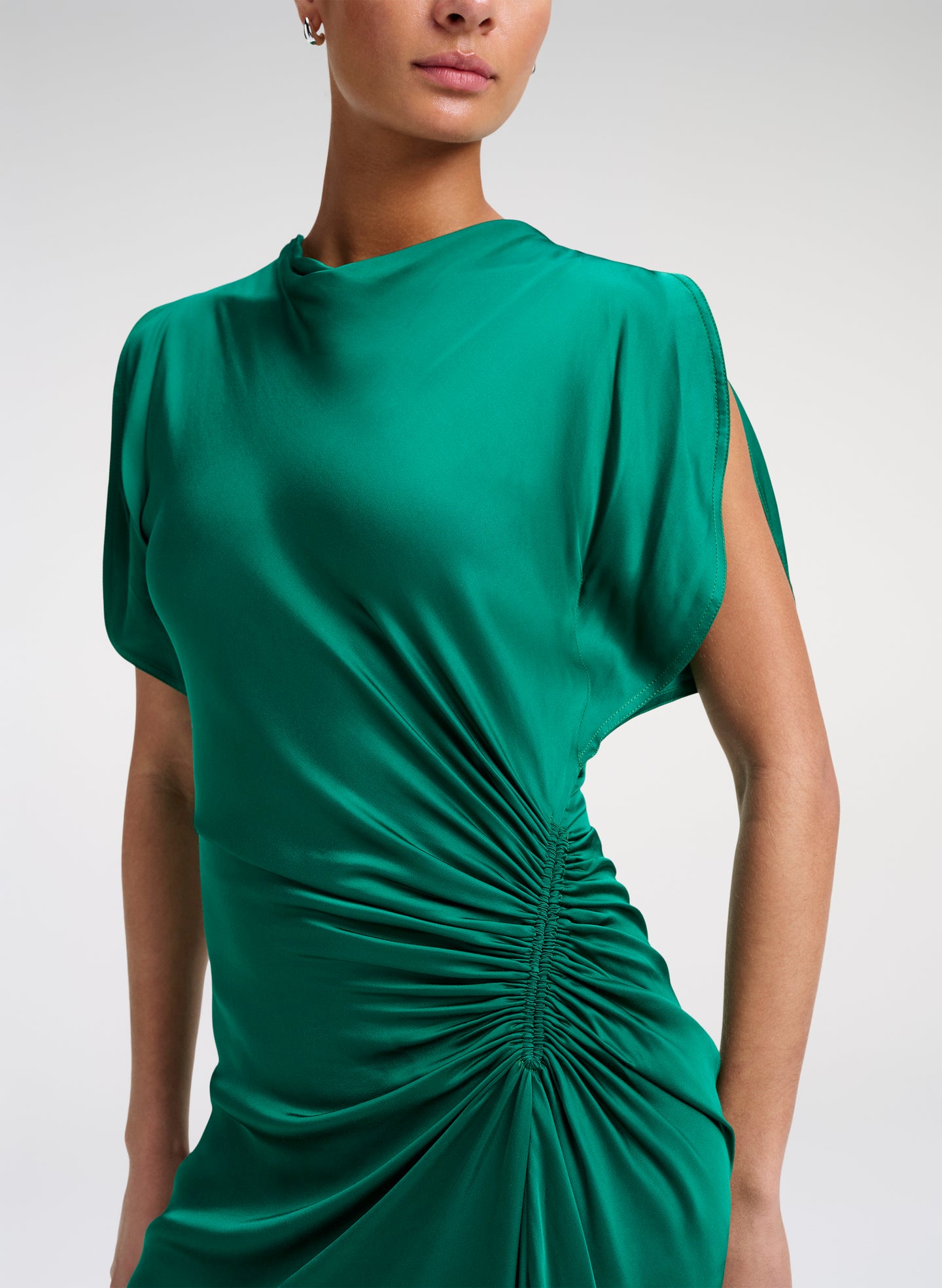 A close-up of a model wearing a green draped dress with short sleeves and a gathered ruched waist detail, set against a plain light gray studio background.