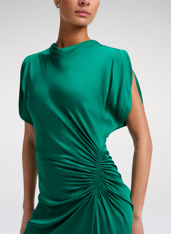 A close-up of a model wearing a green draped dress with short sleeves and a gathered ruched waist detail, set against a plain light gray studio background.