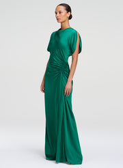 A woman modeling a floor-length emerald green satin gown with draped short sleeves and a gathered ruched waist, photographed against a light gray studio background.