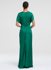 A woman viewed from the back wearing a floor-length emerald green gown with short cap sleeves and a gathered back, photographed against a light gray studio background.