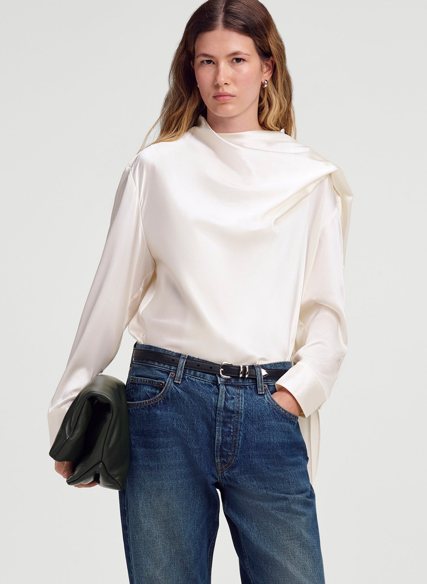A woman modeling a cream-colored draped silk long-sleeve blouse with blue jeans and a black belt, holding a dark green clutch against a light gray studio background.