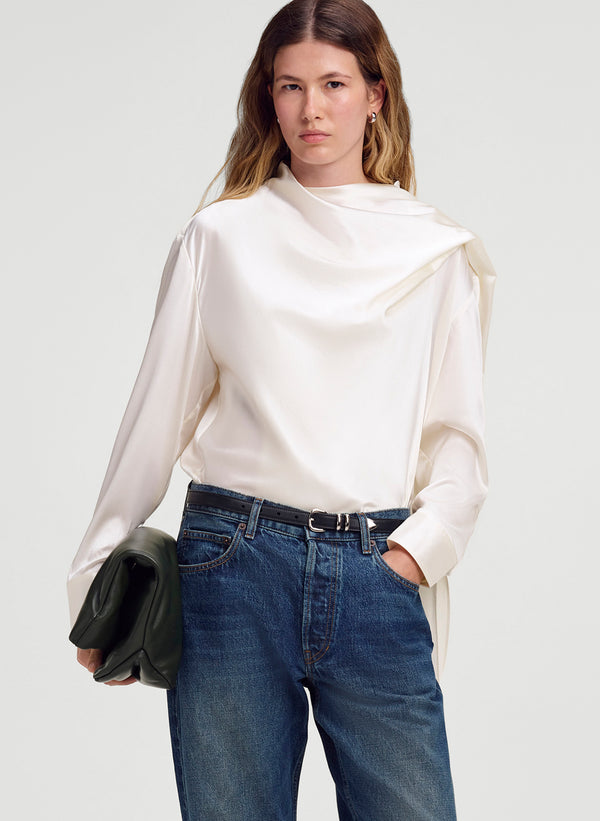 A woman modeling a cream-colored draped silk long-sleeve blouse with blue jeans and a black belt, holding a dark green clutch against a light gray studio background.