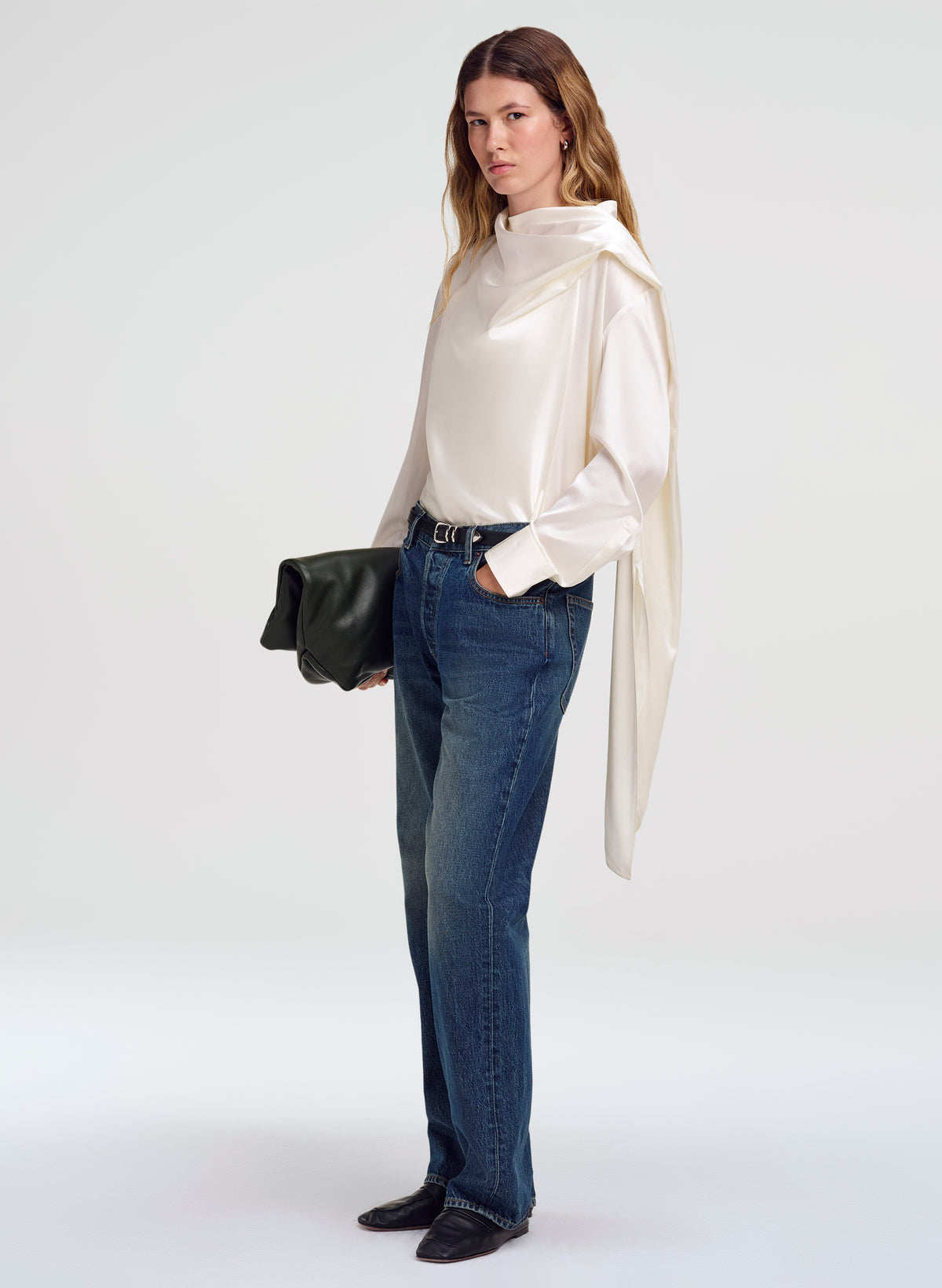 A woman modeling a cream silk long-sleeve blouse with a draped cape-like collar, paired with blue jeans, black shoes, and holding a black clutch bag against a light gray studio background.