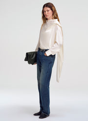 A woman modeling a cream silk long-sleeve blouse with a draped cape-like collar, paired with blue jeans, black shoes, and holding a black clutch bag against a light gray studio background.