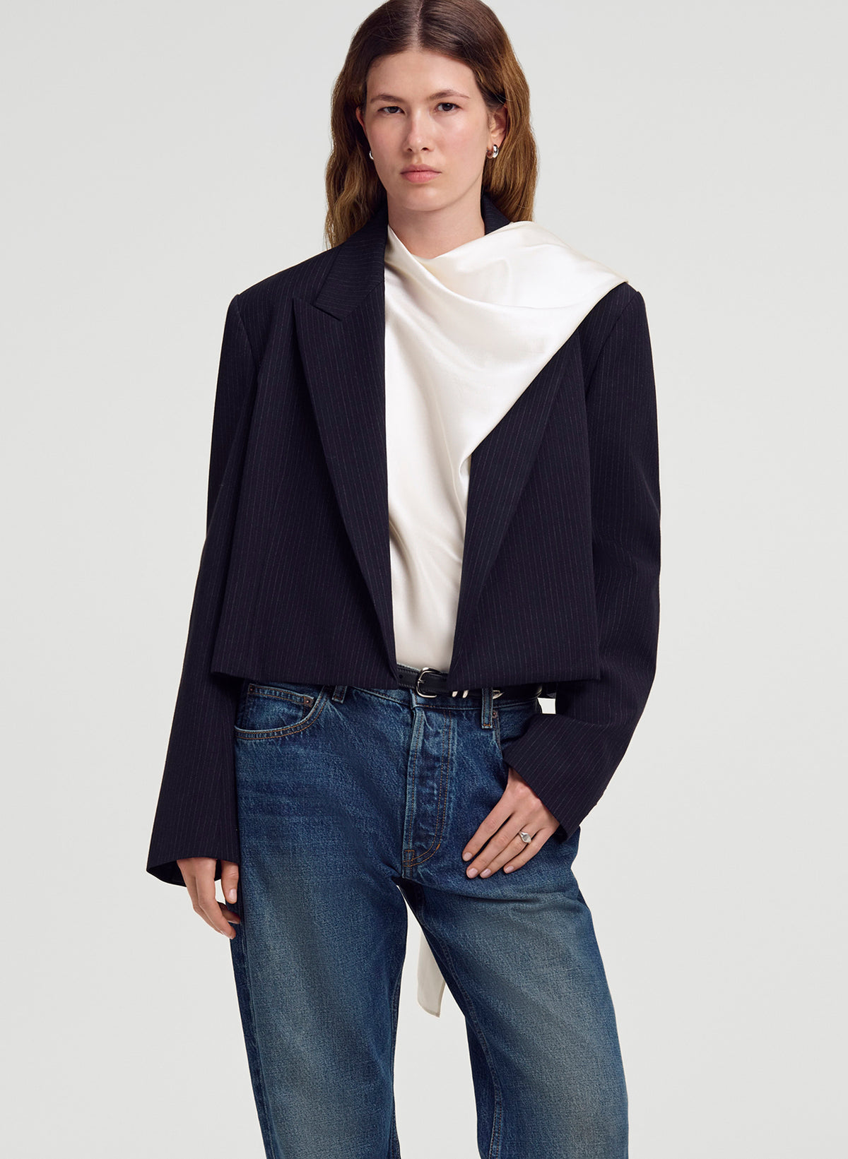 A model wearing a cropped, dark navy blue pinstriped blazer with a unique asymmetrical white draped collar detail, paired with blue jeans and a white top, against a light gray studio background.