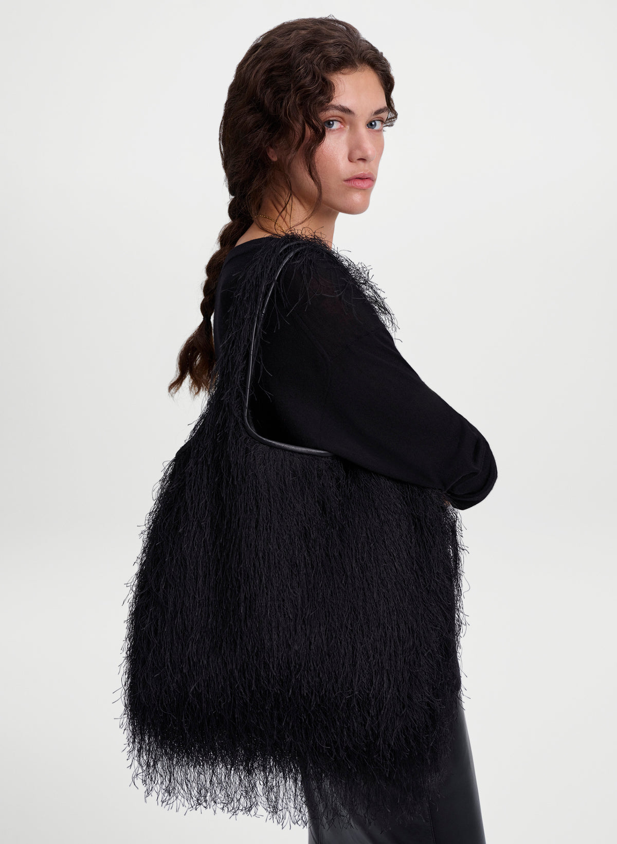 A model with dark brown hair in a side braid is shown from a three-quarters back view, wearing a black long-sleeve top and a large, voluminous black feathered or fringed vest. She is photographed against a clean, light gray studio background.