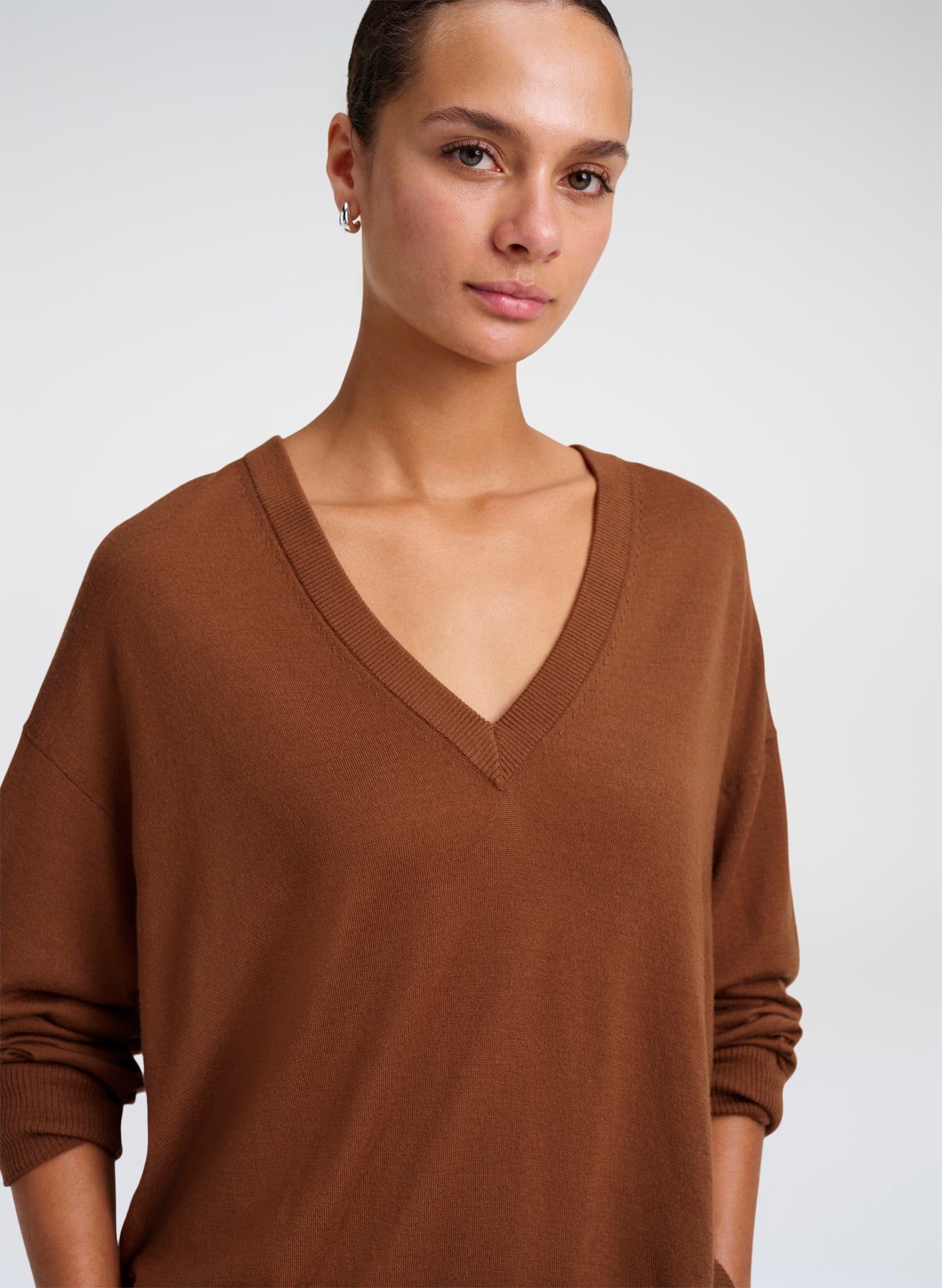 A woman wearing a brown V-neck knit sweater, shown from the chest up against a light gray studio background.