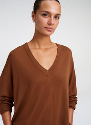 A woman wearing a brown V-neck knit sweater, shown from the chest up against a light gray studio background.