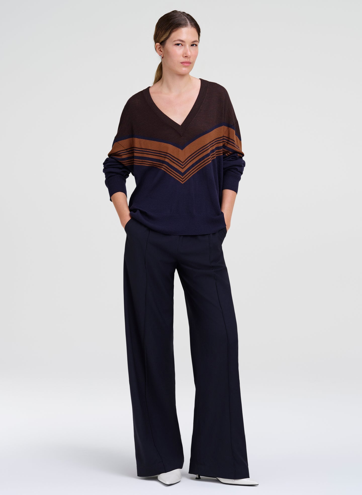 A woman modeling a dark navy blue knit set consisting of a V-neck long-sleeve top with brown chevron stripes on the shoulders and sleeves, paired with matching dark navy wide-leg pants, against a light gray studio background.