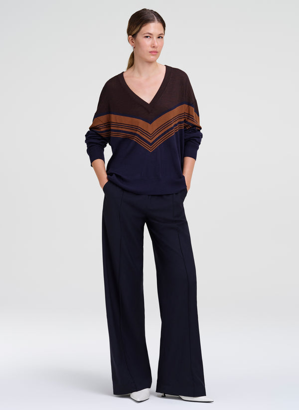 A woman modeling a dark navy blue knit set consisting of a V-neck long-sleeve top with brown chevron stripes on the shoulders and sleeves, paired with matching dark navy wide-leg pants, against a light gray studio background.