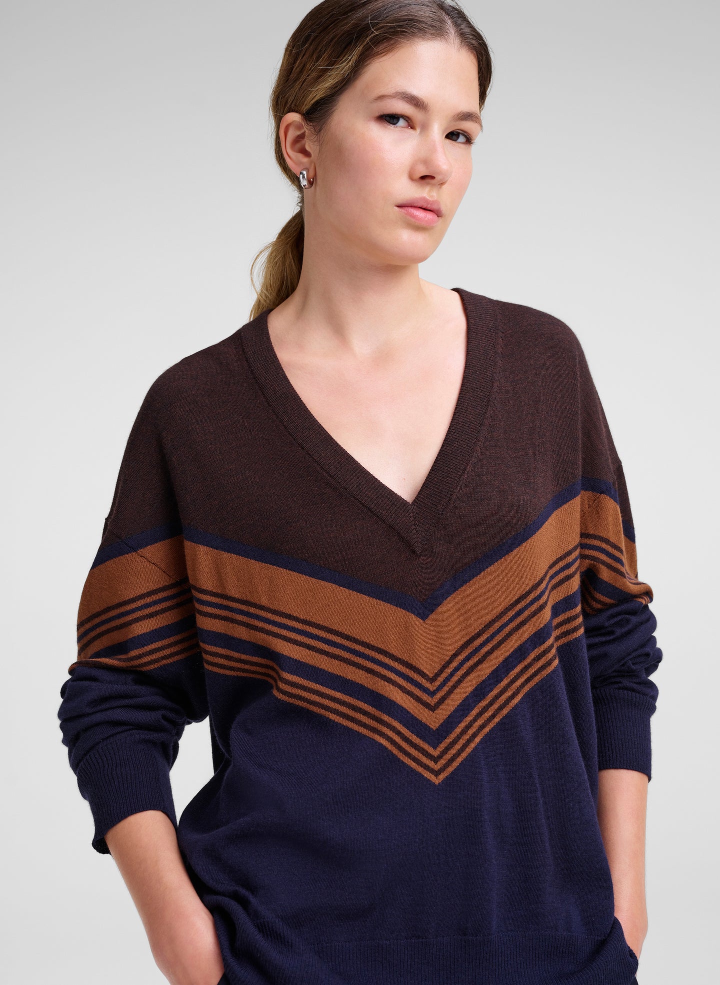 A woman wearing a navy blue and brown v-neck sweater with a chevron stripe pattern across the chest and sleeves, photographed against a light gray studio background.