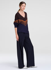 A woman modeling a dark navy blue knit set consisting of a V-neck sweater with brown and gold chevron stripes on the sleeves and chest, paired with matching wide-leg trousers, against a light gray studio background.