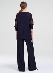 A woman viewed from the back wearing a dark navy long-sleeve top with brown patterned sleeve panels and matching dark wide-leg pants, photographed against a light gray studio background.