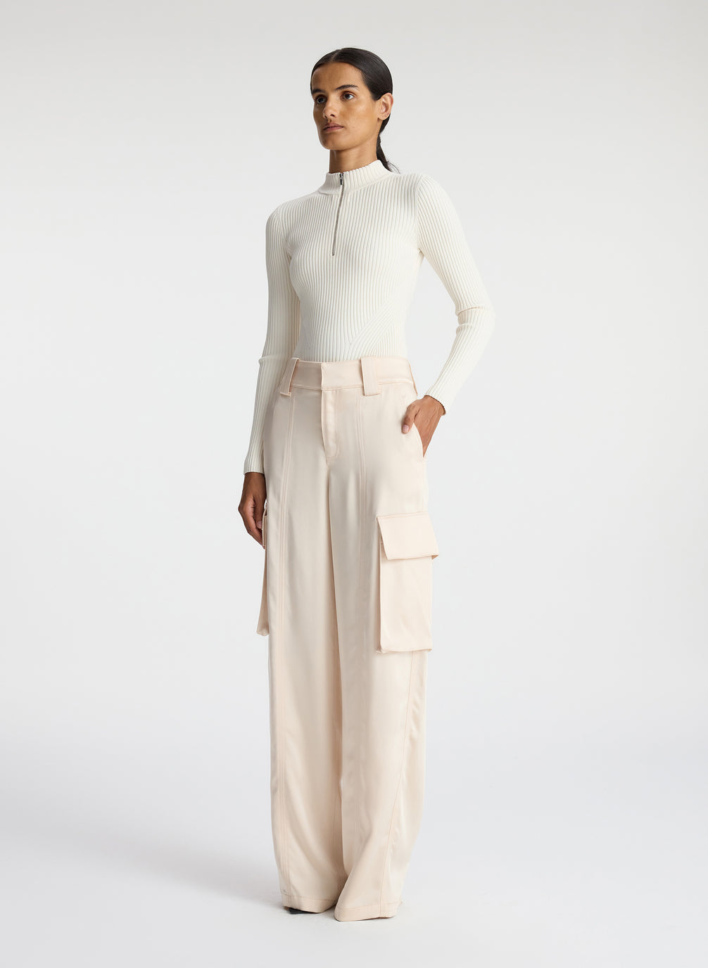 side view of woman wearing white half zip mock neck long sleeve knit top and off white satin cargo pants