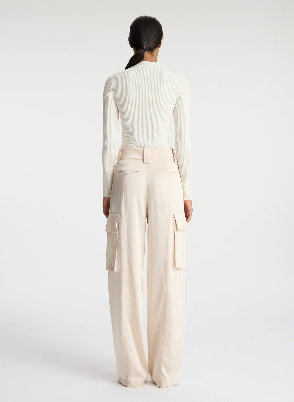 back view of woman wearing white half zip mock neck long sleeve knit top and off white satin cargo pants
