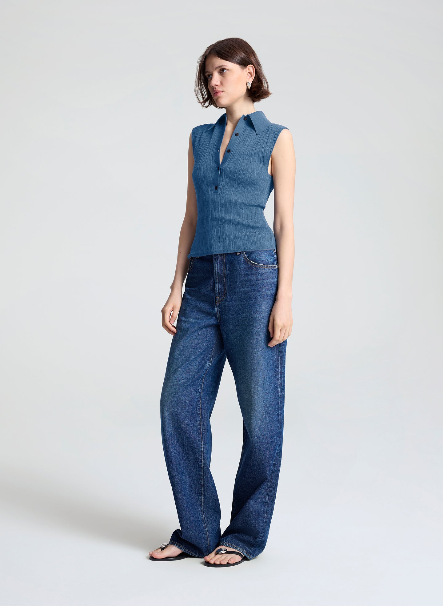 A woman modeling a sleeveless blue knit collared top paired with wide-leg blue jeans and flat sandals, standing against a light gray studio background.