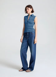 A woman modeling a sleeveless blue knit collared top paired with wide-leg blue jeans and flat sandals, standing against a light gray studio background.