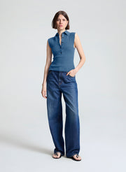 A woman modeling a sleeveless blue collared knit top paired with wide-leg blue jeans and black sandals, standing against a light gray studio background.