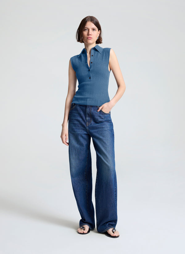 A woman modeling a sleeveless blue collared knit top paired with wide-leg blue jeans and black sandals, standing against a light gray studio background.