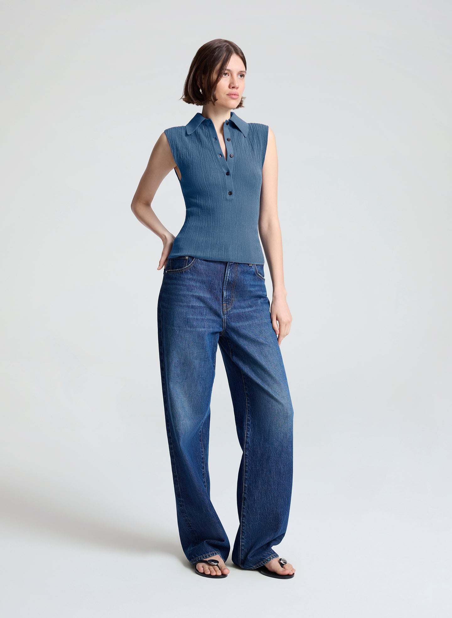 A woman modeling a sleeveless blue button-front top and wide-leg blue jeans against a plain light gray studio background.