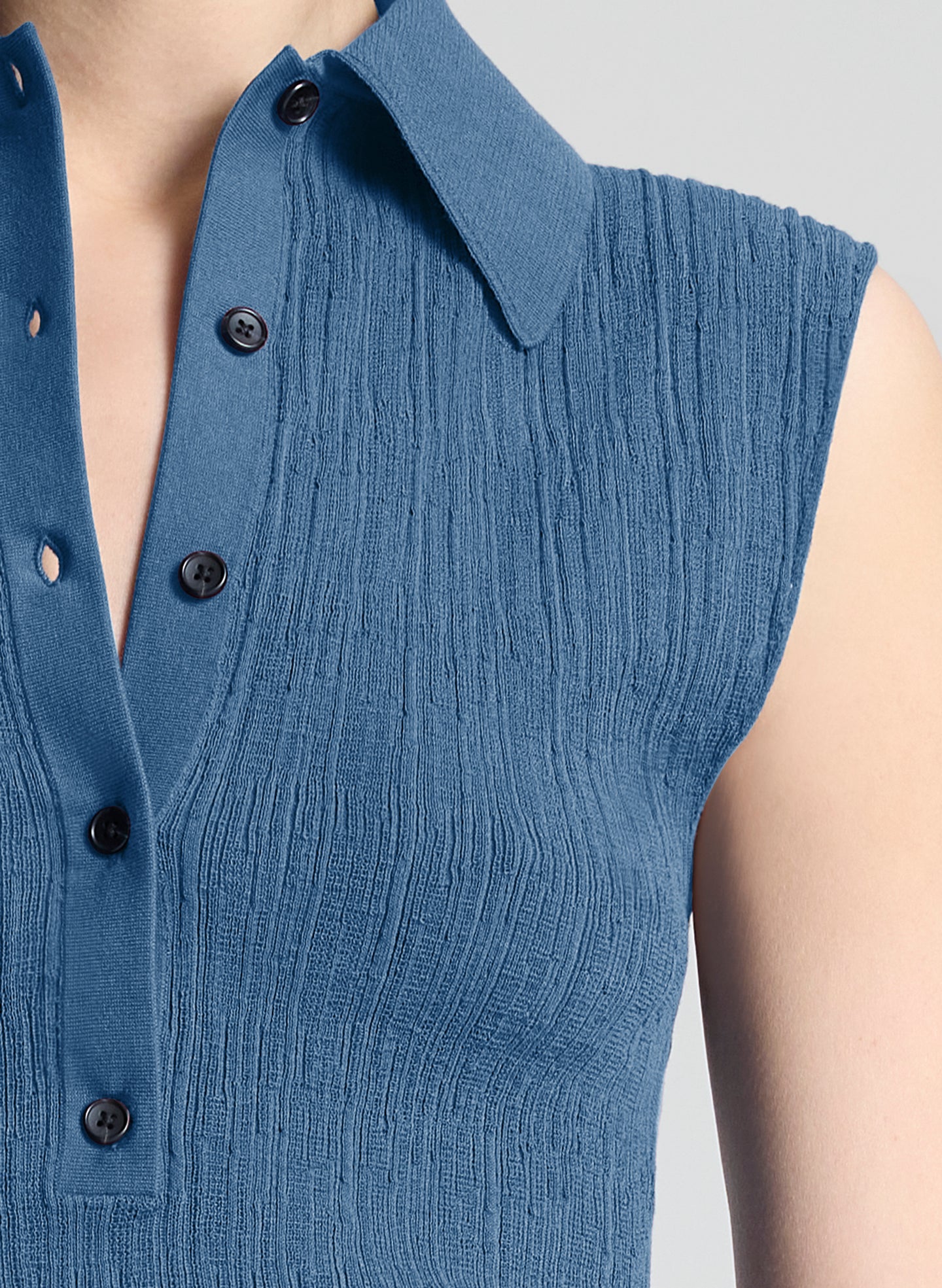 Close-up of a sleeveless blue ribbed knit top with a pointed collar and black buttons, shown on a model against a light gray background.