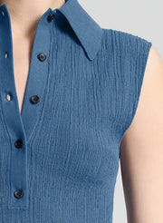 Close-up of a sleeveless blue ribbed knit top with a pointed collar and black buttons, shown on a model against a light gray background.