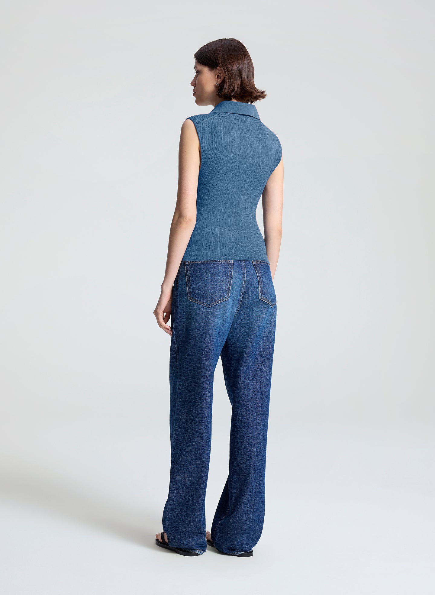 A woman shown from the back wearing a sleeveless blue collared top and wide-leg blue jeans against a plain light gray studio background.