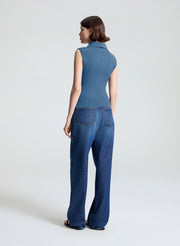 A woman shown from the back wearing a sleeveless blue collared top and wide-leg blue jeans against a plain light gray studio background.