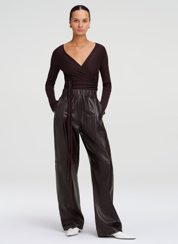 A model wearing a dark brown long-sleeve wrap top and high-waisted brown leather wide-leg trousers, styled with white pointed-toe shoes against a light gray studio background.