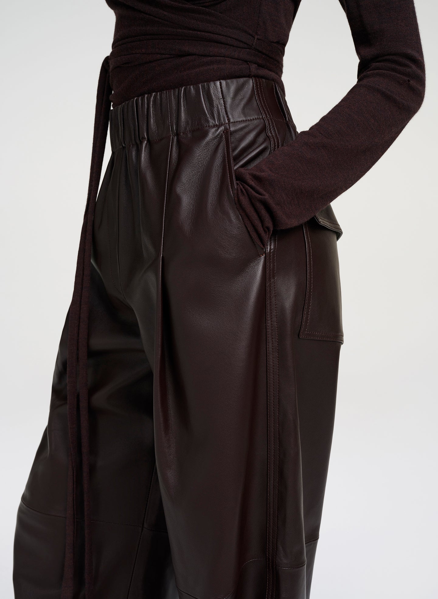 Close-up of a model wearing dark brown leather wide-leg trousers with a matching waist tie and a dark brown long-sleeve top, against a light gray studio background.