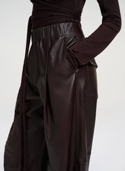 Close-up of a model wearing dark brown leather wide-leg trousers with a matching waist tie and a dark brown long-sleeve top, against a light gray studio background.