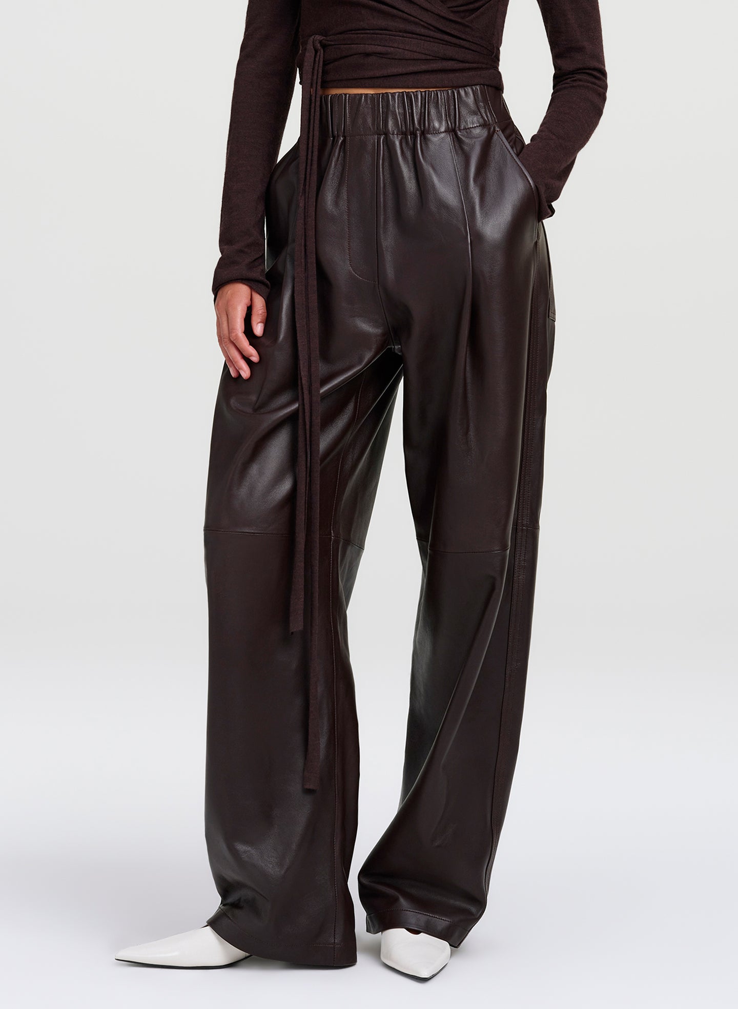 A model wearing dark brown wide-leg  leather pants, paired with a dark brown long-sleeve top and white pointed-toe shoes, shown from the waist down on a light gray studio background.
