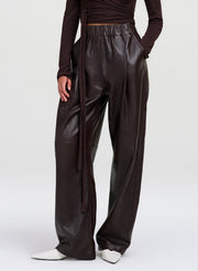 A model wearing dark brown wide-leg  leather pants, paired with a dark brown long-sleeve top and white pointed-toe shoes, shown from the waist down on a light gray studio background.