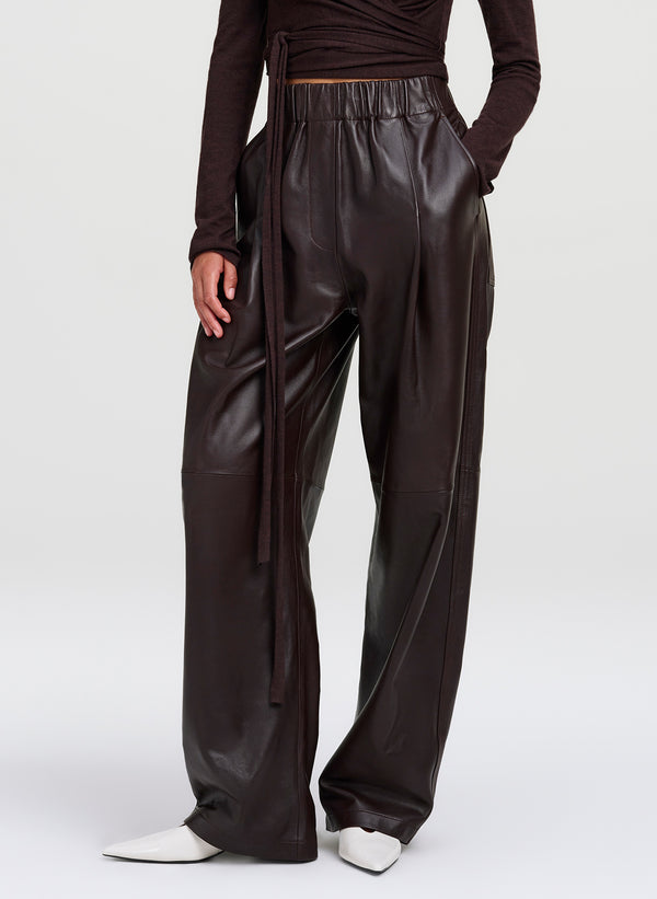 A model wearing dark brown wide-leg  leather pants, paired with a dark brown long-sleeve top and white pointed-toe shoes, shown from the waist down on a light gray studio background.