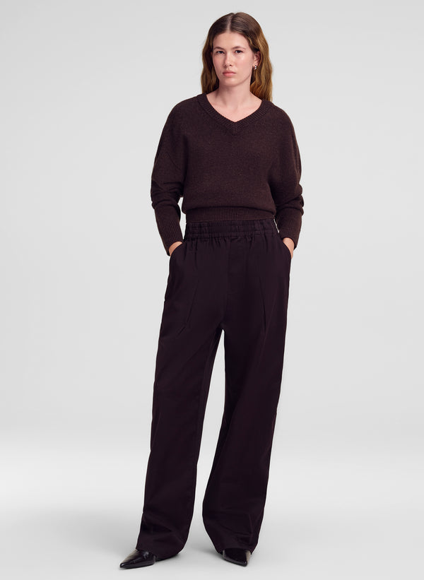 A woman modeling a dark brown v-neck knit sweater and matching dark brown wide-leg trousers against a light gray studio background.